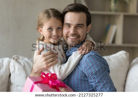 Happy millennial dad embracing beloved daughter kid with love, tenderness, gratitude, holding pink gift wrap, celebrating fathers day, birthday, looking at camera, smiling. Head shot portrait