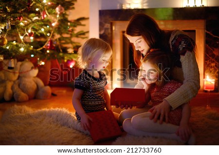 Young mother and her two little daughters opening a magical Christmas gift by a Christmas tree in cozy living room in winter