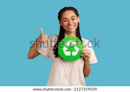 Positive woman with black dreadlocks looking at camera with toothy smile, showing thumb up, holding recycling sign, wearing white shirt. Indoor studio shot isolated on blue background.