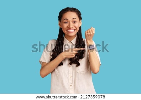 Portrait of happy excited woman with black dreadlocks standing, looking at camera, pointing at her smart watch, wearing white shirt. Indoor studio shot isolated on blue background.