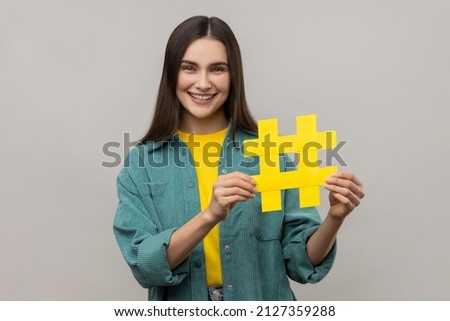 Portrait of happy beautiful woman holding hashtag symbol, promoting viral topic in social network, tagging blog trends, wearing casual style jacket. Indoor studio shot isolated on gray background.