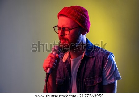 Talented young bearded hipster man singing songs holding microphone, rehearsal before performance, wearing beanie hat and denim vest. Indoor studio shot isolated on colorful neon light background.