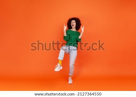 Full length woman with Afro hairstyle wearing green casual style sweater showing rock and roll gesture, standing on one leg, yelling. Indoor studio shot isolated on orange background.