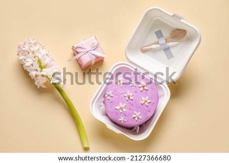 Plastic lunch box with tasty bento cake, gift and flowers on beige background Royalty-Free Stock Photo #2127366680