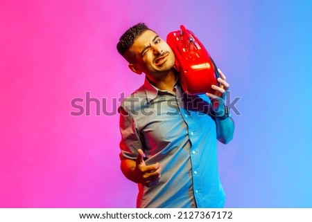 Funny handsome man in shirt listening to music holding red tape recorder on shoulder, winking at camera and showing thumb up. Indoor studio shot isolated on colorful neon light background.