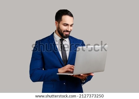 Smiling bearded man holding laptop, typing on keyboard, surfing internet, freelancer working using computer, wearing official style suit. Indoor studio shot isolated on gray background.