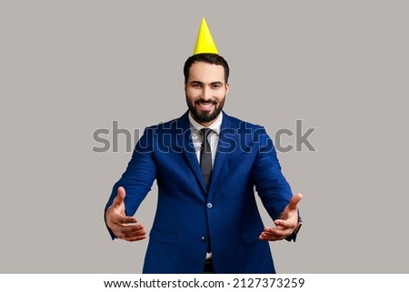 Come into my arms. Friendly smiling man in party cone giving hugs with outstretched hands, welcoming inviting to embrace, wearing official style suit. Indoor studio shot isolated on gray background.