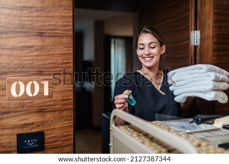 Beautiful young hotel chambermaid in uniform bringing clean towels and other supplies to hotel room. Royalty-Free Stock Photo #2127387344
