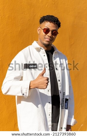smiling latin young man with sunglasses posing looking at the camera and making the I like symbol with his hand, vertical yellow background