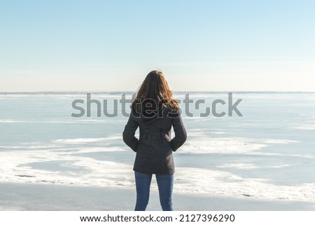 a young woman stands on the shore against the backdrop of a winter and frozen sea. back view Royalty-Free Stock Photo #2127396290