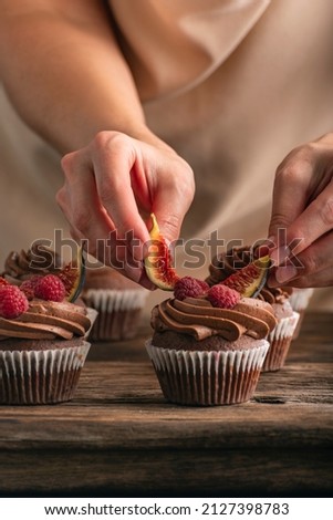Pastry chef decorates muffin with raspberries and figs. Close up photo of cupcakes preparation. Vertical frame. Royalty-Free Stock Photo #2127398783