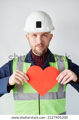National Construction Day concept: cute caucasian bearded construction supervisor, foreman, contractor or engineer in reflective vest and helmet standing by the wall holding red paper heart shape