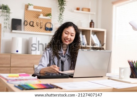 Asian young creative or graphic designer with curly hairstyle drawing or sketching on digital tablet smiling at home.Happiness female freelancer smart and confident enjoying with job or work