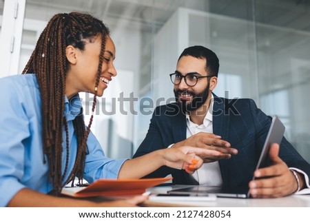 Cheerful multiracial colleagues discussing startup project and smiling during workday in office interior, happy Middle Eastern and African American partners using laptop computer at table desktop Royalty-Free Stock Photo #2127428054