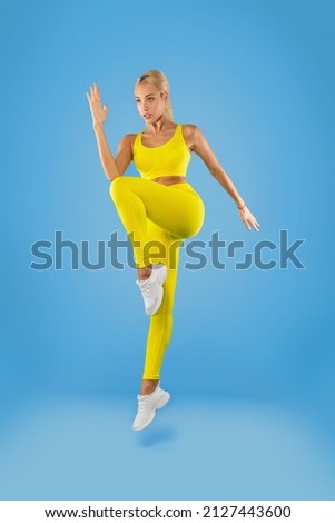 Training Concept. Confident Focused Fit Young Lady In Sportswear Jumping And Lifting Leg Up, Doing High Knee To Elbow Exercise On Blue Studio Wall. Full Body Length Vertical Portrait. Energy And Sport Royalty-Free Stock Photo #2127443600