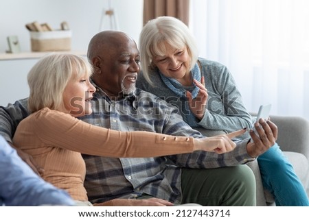 Cheerful multiethnic group of elderly people men and women spending time together at home, sitting on couch and using smartphone, senior black man showing his friends photos on phone, copy space