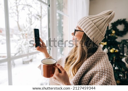 Young caucasian woman standing near window at home and taking photo after snowfall in the morning.