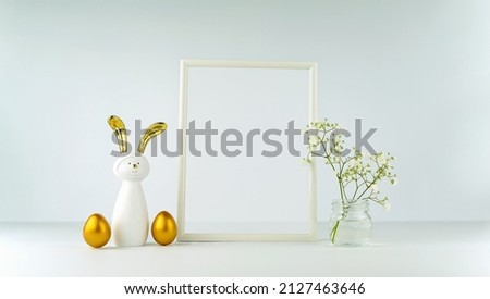 Easter decor on the background of a white table and wall. Eggs and a rabbit with golden ears. Frame and flowers. Minimalist mockup for your text. Banner