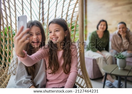 Happy little sisters sitting in wicker rattan hang chair indoors in conservatory and taking selfie.