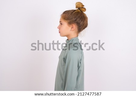 little caucasian kid girl with hair bun wearing technical shirt over white background looking to side, relax profile pose with natural face with confident smile.