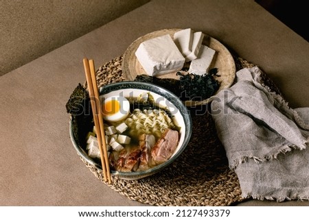 Homemade asian style soup ramen with noodles, grilled duck breast, tofu, seaweed nori chips and boiled egg in ceramic bowl with chopsticks and spoon on brown kitchen table