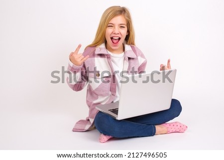 Born to rock this world. Joyful Beautiful caucasian teen girl sitting with laptop in lotus position on white background screaming out loud and showing with raised arms horns or rock gesture.