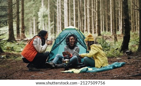 Smiling group of diverse young female friends sitting at their forest campsite drinking coffee and reading a trail map Royalty-Free Stock Photo #2127509756