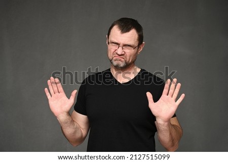 Adult man in a black t-shirt with glasses shows stop with his hands on a gray background