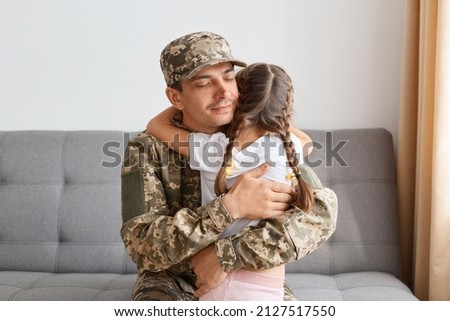 Indoor shot of happy soldier in man camouflage hugging his daughter with closed eyes, missing her kid very much, sitting on sofa indoor at home, arrived home from army.