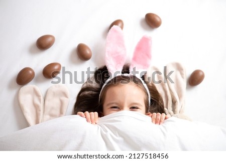 Happy Easter. cute beautiful girl with blue eyes and bunny ears peeking out from under the blanket. Chocolate eggs are scattered on the bed. Egg hunt. Lifestyle High quality photo Royalty-Free Stock Photo #2127518456