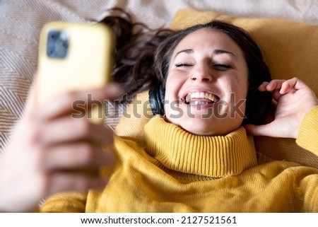 Authentic happy woman listening to music and singing lying on the bed - Beautiful smiling young woman wearing yellow top and holding her smart phone and headphones