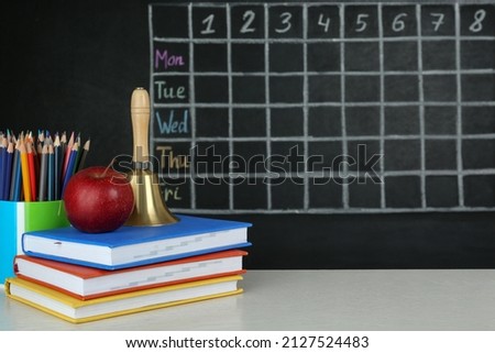 Apple, bell and different stationery on white wooden table near blackboard with chalked school timetable