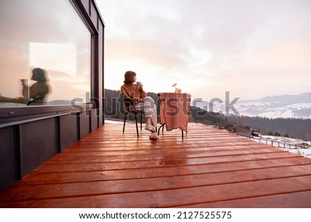 Woman having a breakfast while resting on terrace of tiny house in the mountains, enjoying beautiful landscape during sunrise. Concept of small modern cabins for rest and escape to nature