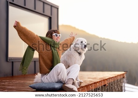 Woman sitting with dog on terrace of tiny house in the mountains enjoying beautiful sunrise landscape. Concept of small modern cabins for rest and escape to nature. Idea of traveling with dog