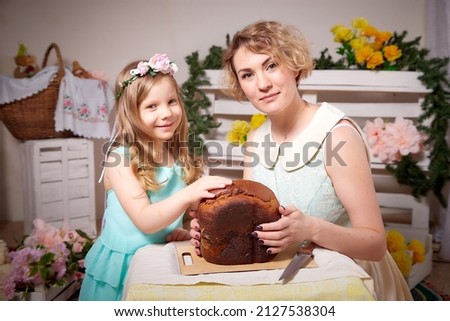Nice mom and beautiful blonde daughter in room durin spring photo shoot before easter with festive bread, kulesh or cake. Happy girl and woman having rest, joy and eating together