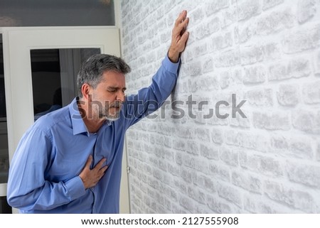 Senior man presses hand to chest has heart attack suffers from unbearable pain closes eyes wears  poses against grey brick wall background. People age and problems with health concept, copy space. Royalty-Free Stock Photo #2127555908