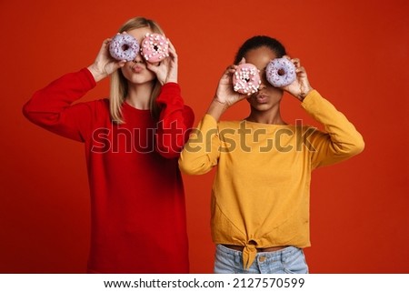 Multiracial two girls grimacing while making fun with donuts isolated over red wall Royalty-Free Stock Photo #2127570599
