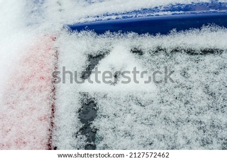 The sign of an apprentice driver on the snow-covered rear window of the automobile. A warning triangular sign with the Russian letter U. Blue car in the winter parking lot.