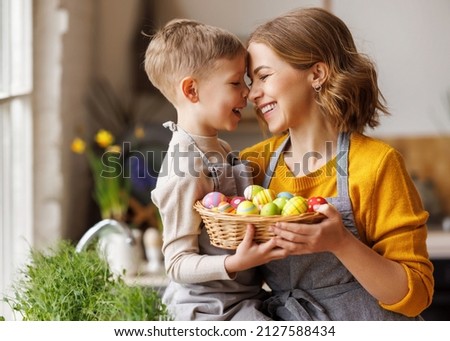 Sweet family portrait of happy mother and little son holding wicker basket full of painted multi-colored Easter eggs, tenderly embracing and smiling in cozy light kitchen at home, selective focus Royalty-Free Stock Photo #2127588434