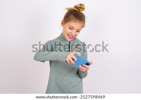 little caucasian kid girl with hair bun wearing technical shirt over white background holding in hands cell playing video games or chatting