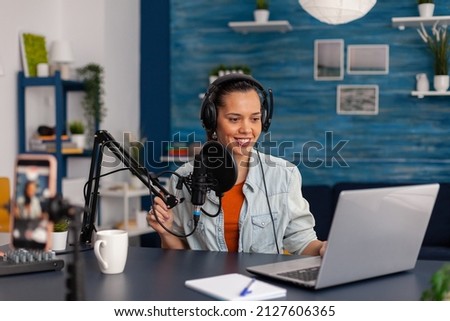 Social media influencer looking at laptop and recording video. Woman vlogger streaming online podcast on smartphone. Content creator in home studio using audio broadcasting equipment.