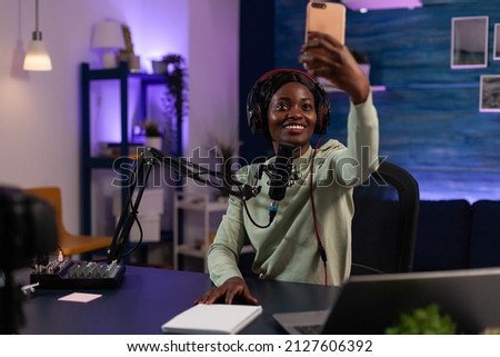 Social media influencer holding smartphone taking selfie while recording vlog content using vlogging production equipment. African american vlogger woman hosting on air talk show