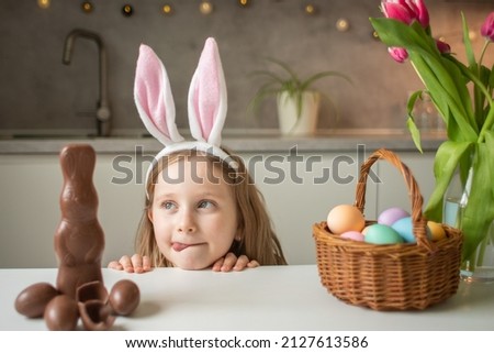 Cute little girl wearing bunny ears eating chocolate Easter rabbit. Kid playing egg hunt on Easter. Adorable child celebrate Easter at home. Royalty-Free Stock Photo #2127613586