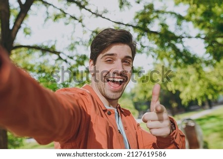 Close up young man in orange jacket walk do selfie shot pov mobile phone point finger camera on you rest relax in spring green city park go down alley sunshine outdoor on nature Urban leisure concept.
