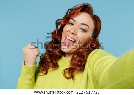 Young close up chubby overweight plus size big fat fit woman in green sweater do selfie shot pov mobile phone winner gesture isolated on plain blue background studio portrait People lifestyle concept