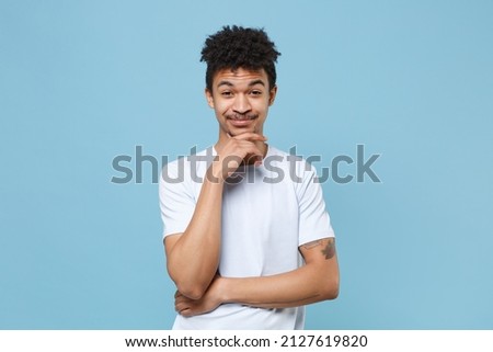 Smiling young african american guy in casual white t-shirt posing isolated on blue background studio portrait. People sincere emotions lifestyle concept. Mock up copy space. Put hand prop up on chin