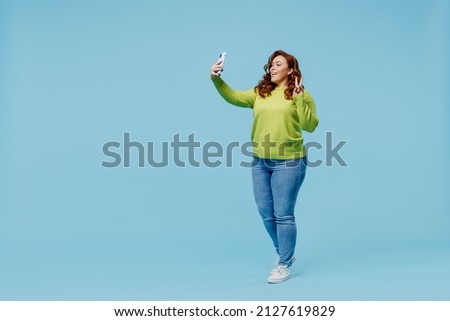 Full body smiling young chubby overweight plus size big fat fit woman wear green sweater doing selfie shot on mobile cell phone show v-sign isolated on plain blue background People lifestyle concept