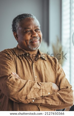 Side view of a happy African retired elderly man standing at home by the window. A satisfied old man looks out the window and smiles as he stands by the window.