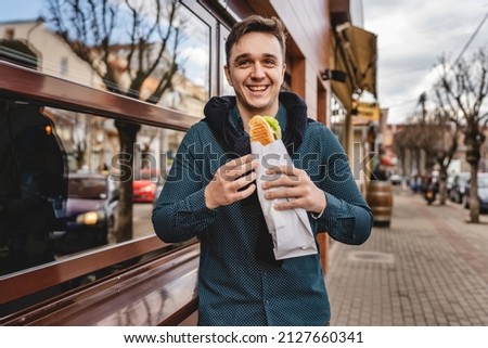 One man standing on the street in autumn spring or winter day holding sandwich wearing jacket eating on the feet while waiting outdoor in front of building real people copy space fast food concept Royalty-Free Stock Photo #2127660341