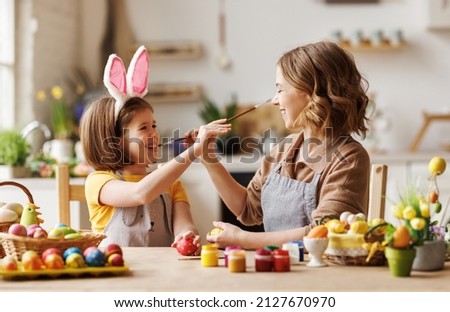 Funny photo of joyful happy family mom and little daughter having fun and fooling around while decorating Easter eggs together for spring holidays, sitting at table in cozy kitchen.  